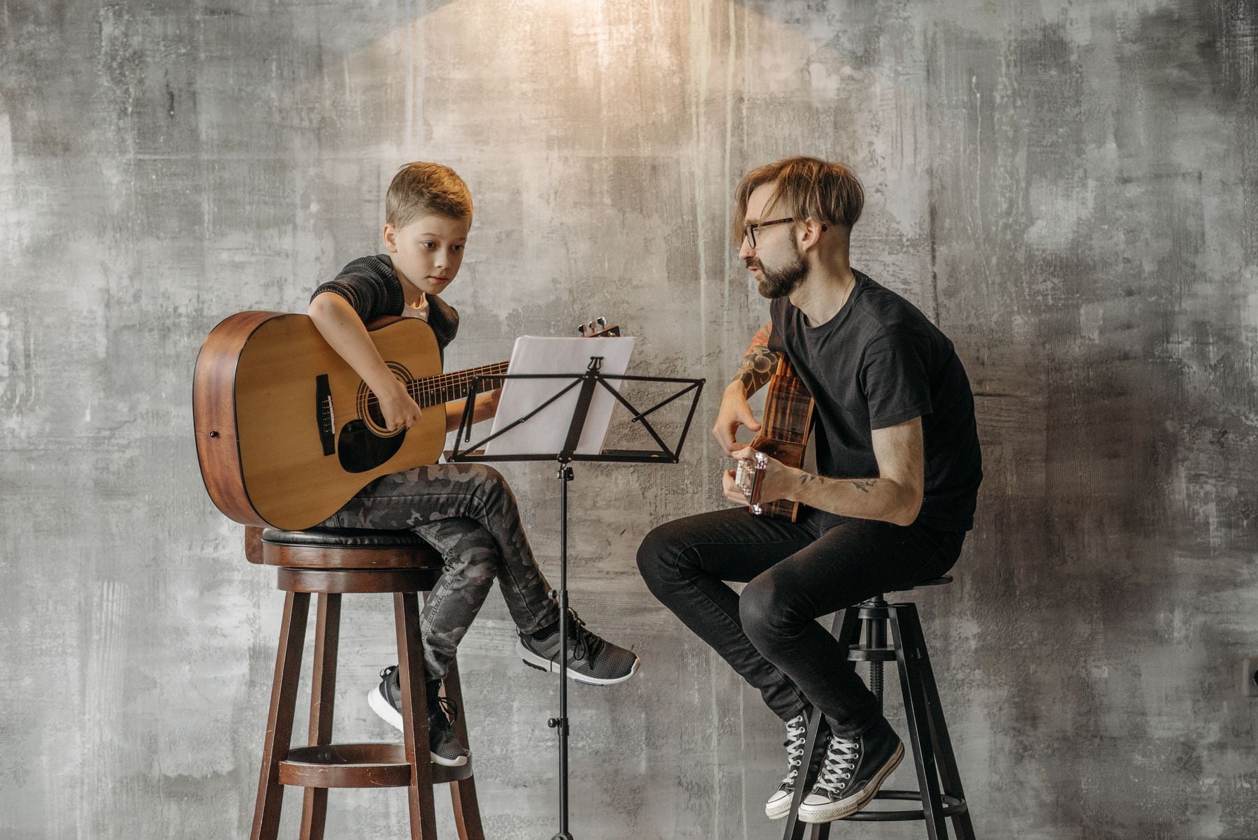 A boy playing an acoustic guitar beside his teacher during a lesson.